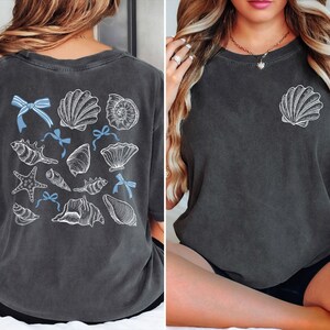 Puede incluir: Camiseta gris oscuro con un dise&ntilde;o de conchas marinas. La parte trasera de la camiseta muestra varias ilustraciones de conchas marinas con lazos azules. Una sola concha est&aacute; en el pecho izquierdo. La camiseta tiene un aspecto vintage.