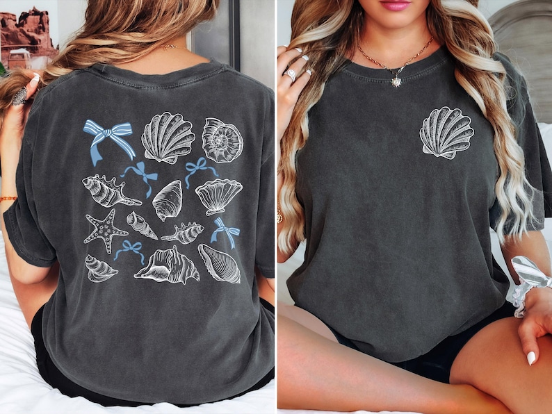 Puede incluir: Camiseta gris oscuro con un dise&ntilde;o de conchas marinas. La parte trasera de la camiseta muestra varias ilustraciones de conchas marinas con lazos azules. Una sola concha est&aacute; en el pecho izquierdo. La camiseta tiene un aspecto vintage.