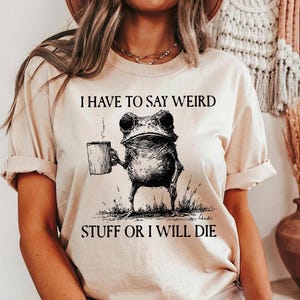 May include: Beige t-shirt with the text "I HAVE TO SAY WEIRD STUFF OR I WILL DIE." The graphic features a detailed illustration of a frog holding a mug. The frog is standing on grass, and the design is in black ink.