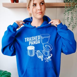 May include: A bright blue hoodie with a white graphic that says "Trashed Panda." The graphic includes a bin, a toilet, a raccoon, and a spilled bottle. The hoodie has a hood and a front pocket.