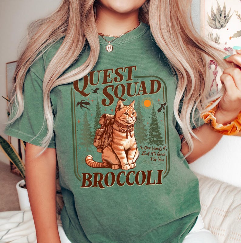May include: Green t-shirt with a retro-style graphic of a cat wearing a backpack and the text "Quest Squad Broccoli" with the tagline "No one wants it, but it's good for you".