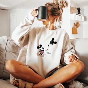 Puede incluir: Una sudadera blanca con un dibujo animado de Mickey Mouse en blanco y negro sosteniendo un globo que dice "30". El texto "Nora" est&aacute; impreso debajo del dibujo animado.
