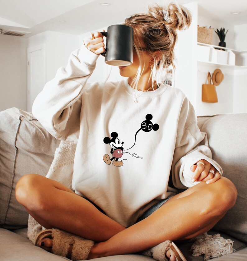 Puede incluir: Una sudadera blanca con un dibujo animado de Mickey Mouse en blanco y negro sosteniendo un globo que dice "30". El texto "Nora" est&aacute; impreso debajo del dibujo animado.