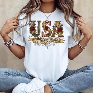 May include: White t-shirt featuring the word "USA" in decorative lettering with floral accents. The "S" is red, and the other letters are gold. A banner below reads "250 YEARS OF FREEDOM".