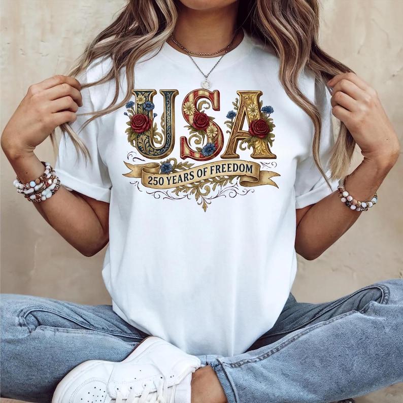 May include: White t-shirt featuring the word "USA" in decorative lettering with floral accents. The "S" is red, and the other letters are gold. A banner below reads "250 YEARS OF FREEDOM".