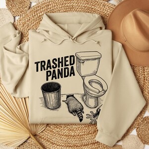 May include: Beige hoodie featuring the text "Trashed Panda" and a black ink graphic of a toilet, bin, and raccoon. A brown hat and woven mats are in the background. The hoodie has a drawstring hood.