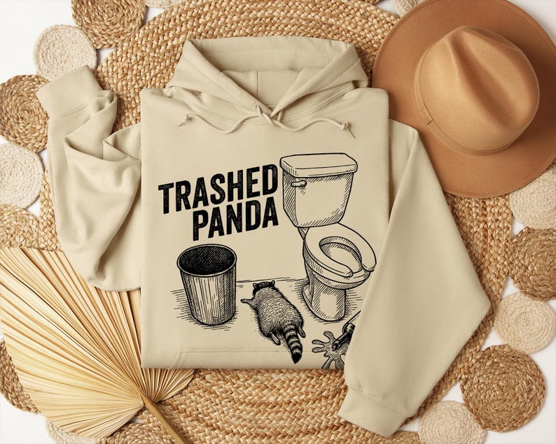 May include: Beige hoodie featuring the text "Trashed Panda" and a black ink graphic of a toilet, bin, and raccoon. A brown hat and woven mats are in the background. The hoodie has a drawstring hood.