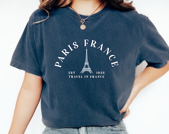 May include: Dark blue t-shirt with the text "PARIS FRANCE" in a semi-circle above an illustration of the Eiffel Tower. Below the tower, the text reads "EST 1928 TRAVEL IN FRANCE". The shirt is a casual style.