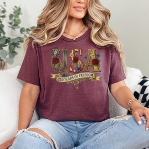 May include: A burgundy t-shirt with the word "USA" in gold lettering, decorated with red roses and blue flowers. Below, a banner reads "250 YEARS OF FREEDOM". The shirt has a V-neck design.