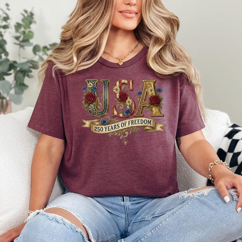 May include: A burgundy t-shirt with the word "USA" in gold lettering, decorated with red roses and blue flowers. Below, a banner reads "250 YEARS OF FREEDOM". The shirt has a V-neck design.