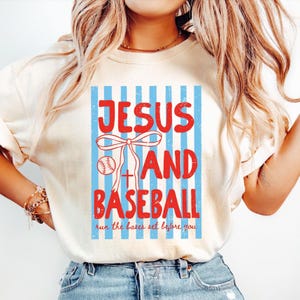May include: Cream-colored t-shirt with a graphic design featuring the words "JESUS AND BASEBALL" in red, set against a blue striped background. A baseball and a cross are incorporated into the design. The phrase "run the bases set before you" is printed at the bottom.