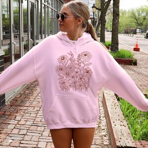 May include: A light pink hoodie with a floral design in shades of brown. The hoodie has a front pocket and a drawstring hood. The floral design features various flowers and leaves. The person in the image is wearing sunglasses.