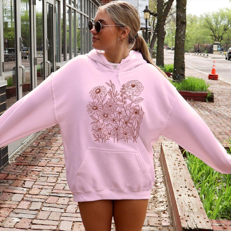 May include: A light pink hoodie with a floral design in shades of brown. The hoodie has a front pocket and a drawstring hood. The floral design features various flowers and leaves. The person in the image is wearing sunglasses.