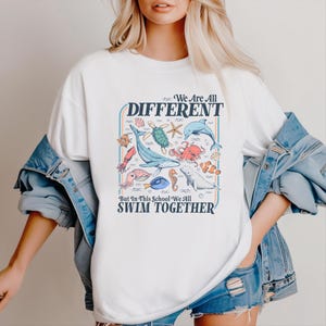 May include: White t-shirt with a colorful graphic of various sea creatures, including a whale, octopus, and seahorse. The text on the shirt reads "We Are All Different, But In This School We All Swim Together."