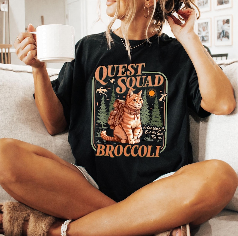 May include: Black t-shirt with a retro-style graphic of a cat wearing a backpack and the text "Quest Squad Broccoli". The text "No one wants it, but it's good for you" is also included in the graphic.