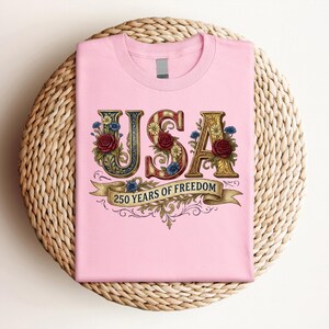 May include: A pale pink t-shirt with the word "USA" in decorative lettering, adorned with floral details. The design incorporates red roses, blue flowers, and a banner reading "250 YEARS OF FREEDOM". The shirt is presented on a woven, circular surface.