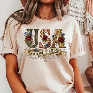 Detailed vintage illustration featuring large ornate gold letters spelling USA intertwined with red roses, blue cornflowers, and white daisies. A beige ribbon banner at the bottom reads 250 YEARS OF FREEDOM in dark classic typography.