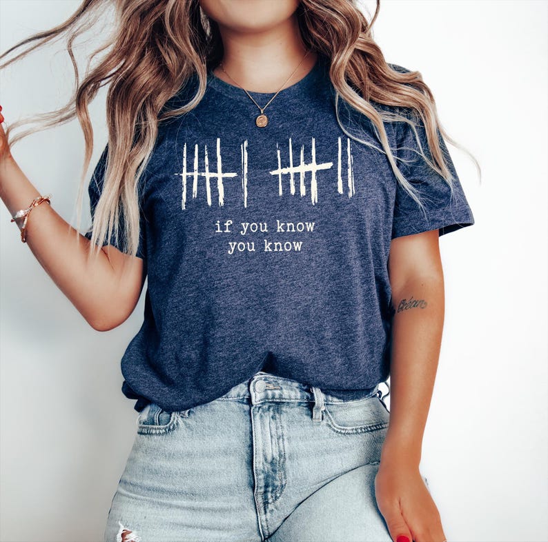 May include: Navy blue t-shirt with white graphic lines and the text "if you know you know". The shirt has a crew neck and short sleeves. The model is wearing light blue denim jeans.