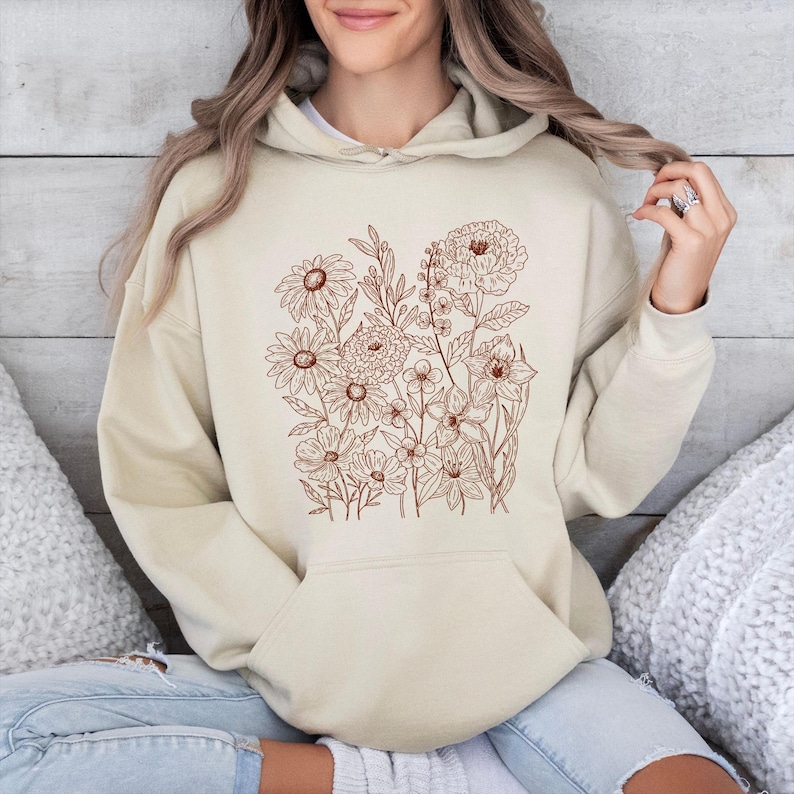 May include: A beige hoodie featuring a brown floral design on the front. The design includes various flowers and leaves. The hoodie has a hood and a front pocket. The model is wearing the hoodie.