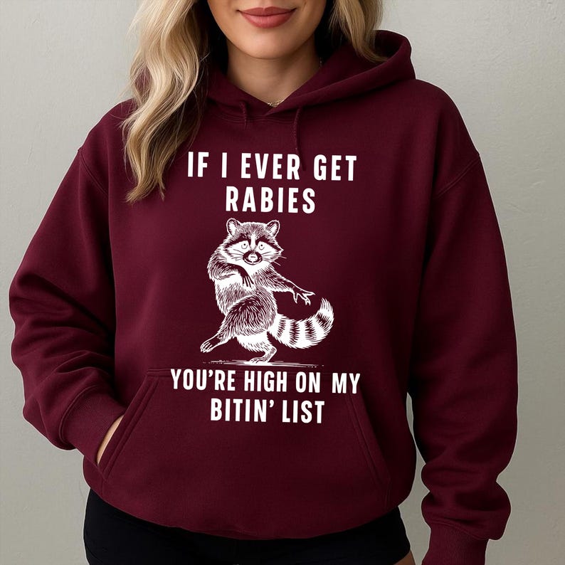 Op de afbeelding: Bordeaux hoodie met een witte afbeelding van een wasbeer en de tekst "IF I EVER GET RABIES YOU'RE HIGH ON MY BITIN' LIST". De hoodie heeft een voorzak en een capuchon met trekkoord. De wasbeer-afbeelding is gedetailleerd met zwart-witte vacht.