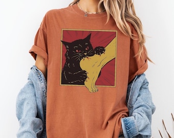 May include: Burnt orange t-shirt featuring a graphic of a black cat biting a foot. The cat has red eyes and is set against a red and yellow background. The shirt has short sleeves and a crew neck.