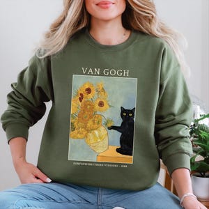 May include: Olive green sweatshirt featuring a print of Van Gogh's "Sunflowers (Third Version)" painting. The artwork includes a vase of sunflowers and a black cat. The text "VAN GOGH" and "SUNFLOWERS (THIRD VERSION) - 1888" are also printed on the sweatshirt.