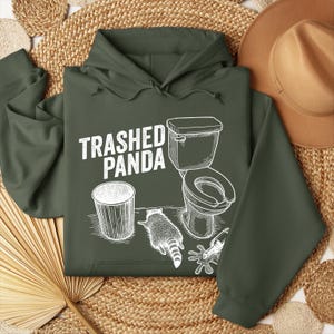 May include: Dark green hoodie featuring the text "Trashed Panda" and a white graphic of a raccoon, toilet, and bin. The design contrasts with the hoodie's colour. The hoodie includes a hood and drawstrings.