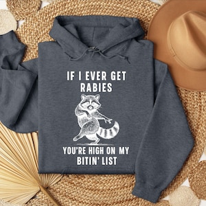 Op de afbeelding: Donkergrijze hoodie met de tekst "IF I EVER GET RABIES" en "YOU'RE HIGH ON MY BITIN' LIST" in het wit. Een witte cartoon wasbeer staat onder de tekst. De hoodie heeft een capuchon met trekkoord en lange mouwen.