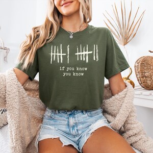 May include: Olive green t-shirt with white vertical line graphics and the text "if you know you know". The shirt is a short-sleeved crew neck style.
