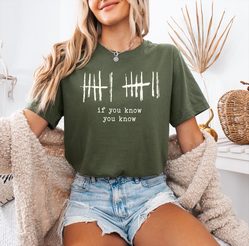 May include: Olive green t-shirt with white vertical line graphics and the text "if you know you know". The shirt is a short-sleeved crew neck style.
