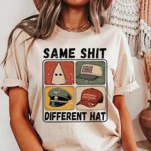 May include: Beige t-shirt with the text "SAME SHIT DIFFERENT HAT" above a graphic of four hats: a white hood, a Confederate flag hat, a military hat, and a red "Make America Great Again" hat.