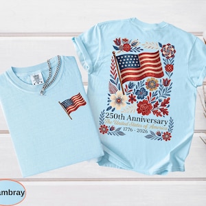 May include: Two light blue t-shirts. One has a small American flag embroidered on the chest. The other has a large American flag design on the back, surrounded by flowers and the text "250th Anniversary The United States of America 1776-2026."