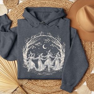 May include: Dark blue hoodie featuring a white graphic of four witches dancing in a circle under a crescent moon and stars. The design is surrounded by a tree branch border. The hoodie has a hood and a kangaroo pocket.