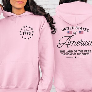 Pu&ograve; includere: Una felpa con cappuccio rosa chiaro con tasca frontale e cappuccio. La parte anteriore presenta la scritta "Since 1776" all'interno di un cerchio di stelle. Il retro mostra "United States of America" con due bandiere americane e le frasi "The Land of the Free" e "The Home of the Brave."