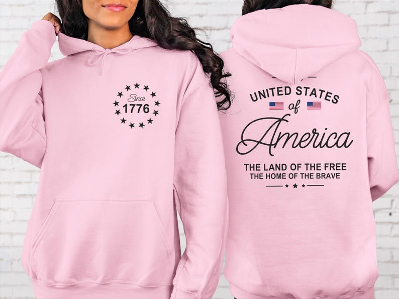 Pu&ograve; includere: Una felpa con cappuccio rosa chiaro con tasca frontale e cappuccio. La parte anteriore presenta la scritta "Since 1776" all'interno di un cerchio di stelle. Il retro mostra "United States of America" con due bandiere americane e le frasi "The Land of the Free" e "The Home of the Brave."