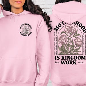 May include: Light pink hoodie with the text "MOTHERHOOD IS KINGDOM WORK" and a floral design. The back of the hoodie has the same text and design, along with a Bible verse reference. A comfortable and stylish sweatshirt.