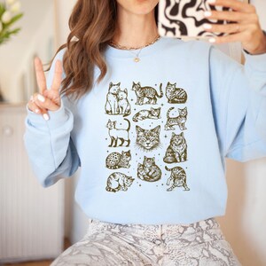 May include: Light blue sweatshirt featuring a grid of various cat illustrations in a sepia tone. The cats are depicted in different poses and styles, creating a charming and whimsical design. The sweatshirt is a comfortable, casual style.