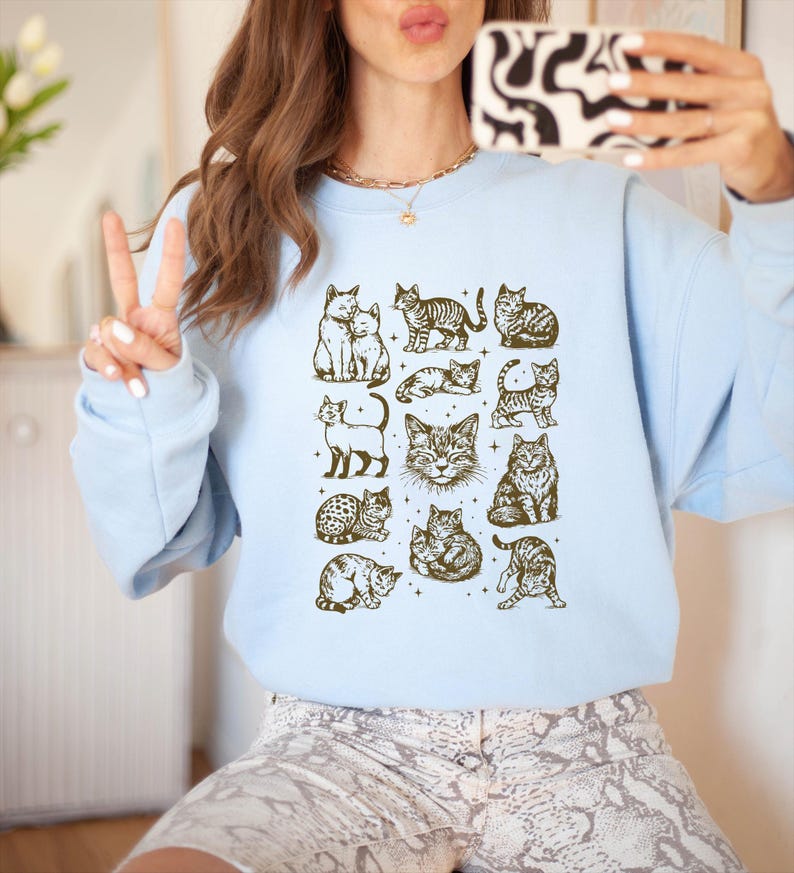 May include: Light blue sweatshirt featuring a grid of various cat illustrations in a sepia tone. The cats are depicted in different poses and styles, creating a charming and whimsical design. The sweatshirt is a comfortable, casual style.