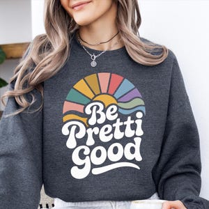 May include: Dark gray sweatshirt with a retro design featuring a colorful sunburst and the words "Be Pretti Good" in white. The design includes shades of yellow, orange, red, teal, and purple. The sweatshirt is a casual style.