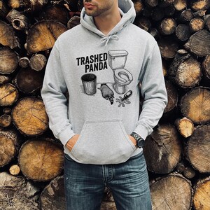 May include: A light grey hooded sweatshirt with a front pocket and a drawstring hood. The graphic on the front displays the text "Trashed Panda" above a drawing of a toilet, bin, raccoon, and broken bottle.
