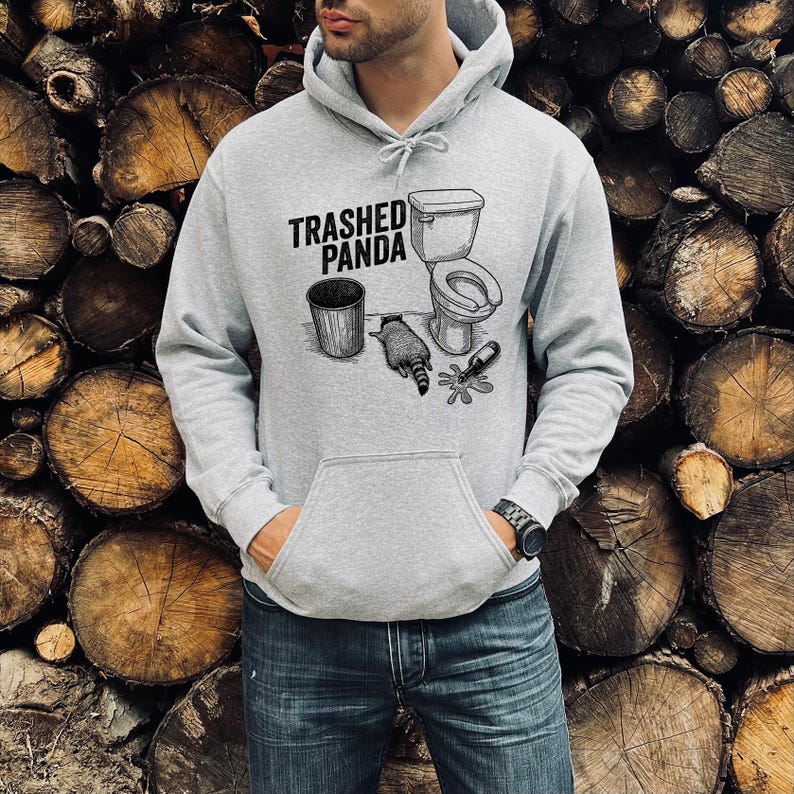 May include: A light grey hooded sweatshirt with a front pocket and a drawstring hood. The graphic on the front displays the text "Trashed Panda" above a drawing of a toilet, bin, raccoon, and broken bottle.