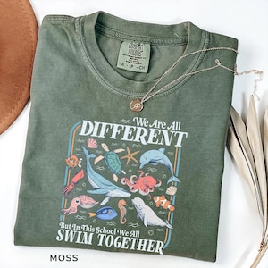 May include: Moss green t-shirt with the text "We Are All Different But In This School We All Swim Together" and various sea creatures. A gold necklace with a pendant rests on the shirt.