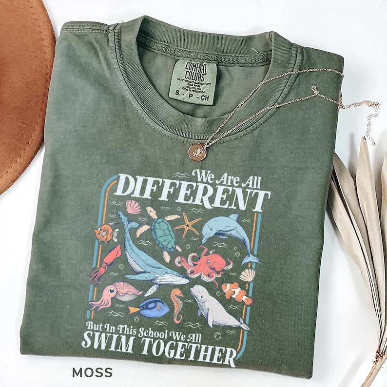 May include: Moss green t-shirt with the text "We Are All Different But In This School We All Swim Together" and various sea creatures. A gold necklace with a pendant rests on the shirt.