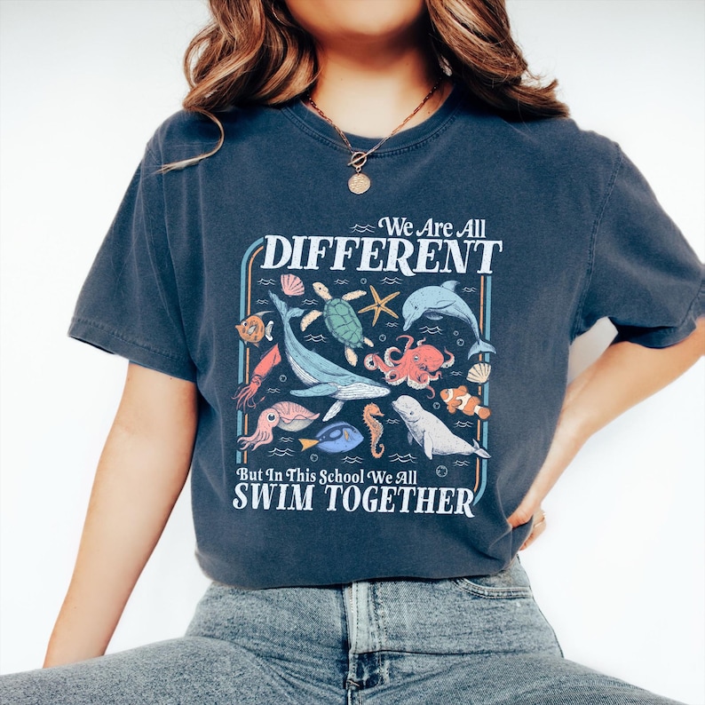 May include: Dark blue t-shirt with a colorful graphic of various sea creatures, including a whale, octopus, and seahorse. The text on the shirt reads "We Are All DIFFERENT But In This School We All SWIM TOGETHER."
