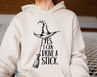 May include: A cream-colored hoodie with a black graphic of a witch's hat and broom. The text "YES I CAN DRIVE A STICK" is printed below the hat. The hoodie has a front pocket and a hood.
