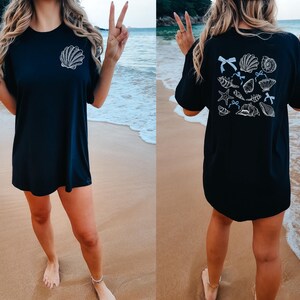 Puede incluir: Camiseta negra con un dise&ntilde;o de concha blanca en la parte delantera y una colecci&oacute;n de conchas en la parte trasera. La camiseta se usa en una playa con el oc&eacute;ano de fondo.