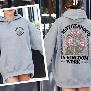 May include: A heather gray hoodie with the text "MOTHERHOOD IS KINGDOM WORK" and a floral design. The back of the hoodie features the same text and design, along with a Bible verse. A comfortable, casual sweatshirt.