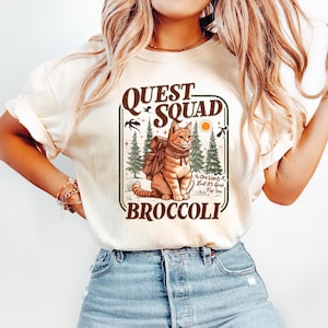 May include: A white t-shirt with a graphic of a cat wearing a backpack and the text "QUEST SQUAD BROCCOLI" in brown lettering. The cat is surrounded by trees and the text "No one wants it, but it's good for you".