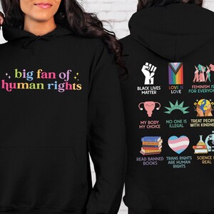 May include: Black hoodie with the text "big fan of human rights" in colorful letters on the front. The back features various graphics and phrases, including "Black Lives Matter," "Love is Love," and "Trans Rights are Human Rights."