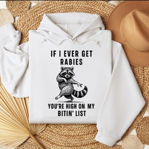 Op de afbeelding: Witte hoodie met een wasbeer-afbeelding en de tekst "IF I EVER GET RABIES YOU'RE HIGH ON MY BITIN' LIST". De wasbeer is afgebeeld in zwart en wit. De hoodie heeft een capuchon met trekkoord.